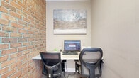 This single-floor space offers a compelling contrast between raw architectural character and clean, contemporary finishes. The most distinctive feature is a full exposed brick wall, displaying warm red and orange tones with light-colored mortar that adds texture and visual depth throughout the interior. This element anchors the space with an industrial-meets-refined aesthetic.

The entry area is bright and welcoming, finished with light gray tile flooring and framed by large windows fitted with white horizontal blinds that allow natural light to filter through. A white paneled door with glass inserts provides views to an outdoor walkway, while a warm wood-framed glass door nearby adds a classic architectural touch, creating an inviting transition into the space.

Adjacent to the entry, a compact but well-organized workstation area is tucked against the exposed brick wall. A shared desk accommodates a desktop computer setup and two ergonomic office chairs, making it a functional workspace for one or two users. A large abstract painting in muted gold, silver, and teal tones hangs above the desk, adding an artistic focal point.

The remaining walls feature a smooth, warm beige finish with crisp white trim, contributing to a calm and neutral overall ambiance that balances the boldness of the brick.
