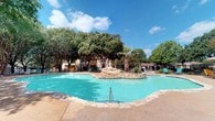 This single-story amenity space features two distinct areas designed for recreation and wellness. The outdoor pool area is a standout highlight, showcasing a generously sized, freeform swimming pool with vivid turquoise water and a decorative stone waterfall centerpiece. The pool is bordered by a concrete deck finished with stone edging, providing space for lounge seating. Mature trees, including a prominent palm, frame the area, creating a shaded, resort-like atmosphere under open skies. A perimeter fence encloses the space, offering privacy and security.

The fitness center is bright and thoughtfully designed, featuring bold teal, orange, and neutral wall color-blocking that lends a vibrant, energetic ambiance. Large arched windows allow generous natural light to fill the room while offering views of the surrounding landscape. Wood-look flooring runs throughout, and white decorative columns add architectural character. Equipment includes treadmills, an elliptical machine, stability balls on a dedicated rack, free weights, and a balance scale. Mirrored walls enhance the sense of space, and French doors provide convenient access to adjacent areas of the property.