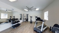 This dedicated fitness center offers a well-organized and fully equipped workout environment designed to support a wide range of exercise routines. The space features durable wood-look flooring throughout, providing both a polished aesthetic and a resilient surface suited for high-activity use. Bright white walls and recessed lighting panels contribute to a clean, energizing atmosphere, while a large ceiling fan with an extended blade span promotes steady air circulation during workouts. Natural light filters in through a window, further enhancing the open and airy feel of the room. A full-length wall mirror along one side visually expands the space and allows users to monitor their form during exercise. The equipment layout is thoughtfully arranged, featuring a multi-station cable machine for strength training, a stationary bike and treadmill for cardio sessions, and a well-stocked dumbbell rack offering a range of free weights. The overall design balances functionality and comfort, making efficient use of the available floor space without feeling crowded. The space is clean and well-maintained, reflecting a commitment to providing a quality fitness experience.