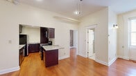 Located at 20 St. Louis Pl in Buffalo, NY, this efficiently designed single-floor unit offers a thoughtful layout comprising a living room, kitchen, bathroom, and an additional versatile room. Warm hardwood flooring flows throughout, complementing the soaring ceilings that lend a sense of openness well beyond the unit's compact footprint. The kitchen is a standout feature, showcasing rich dark cabinetry, granite countertops, stainless steel appliances including a refrigerator and gas range, a central island for additional prep space, and a stacked in-unit washer and dryer tucked neatly within the space. The living area is anchored by a ceiling fan with light fixture and benefits from natural light streaming through windows. A secondary room impresses with dramatic exposed brick walls spanning floor to ceiling, adding historic character and texture, along with a window that invites in natural light. The bathroom is finished with tile flooring, a glass-enclosed shower, and a glass-block window that provides privacy while still allowing light. Crisp white trim and neutral walls throughout create a clean, cohesive aesthetic that highlights the space's blend of historic charm and modern updates.