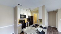 This single-floor space centers around a well-appointed kitchen that balances practicality with modern aesthetics. The kitchen is outfitted with sleek black appliances, including a refrigerator, range, and built-in microwave, all complemented by crisp white cabinetry that extends to the ceiling, maximizing storage capacity. A tasteful tile backsplash adds texture and visual interest behind the cooking area.

The granite countertops are a standout feature, offering generous preparation space with a distinctive speckled pattern. A peninsula extends the counter space into a breakfast bar, complete with a undermount sink and an arched gooseneck faucet, making it an ideal spot for casual dining or entertaining. The bar seating area is set with place settings, highlighting its social functionality.

Track lighting mounted on the ceiling provides targeted illumination throughout the kitchen. The flooring transitions to dark hardwood, grounding the space with warmth. A glimpse through the hallway reveals a tiled bathroom, suggesting a thoughtfully connected layout throughout the residence. Overall, the space presents a clean, contemporary design with cohesive finishes.