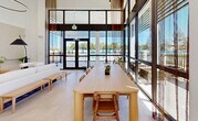 Located in Homestead, Florida, this single-floor community clubhouse offers a thoughtfully designed amenity space that combines modern aesthetics with functional versatility. The interior is characterized by soaring ceilings, crisp white walls, and large floor-to-ceiling windows that flood the space with natural light and frame views of the outdoor pool area beyond.

The lounge and social area anchors the space with a sophisticated, contemporary feel. A plush white sectional sofa, warm-toned accent seating, and a circular pendant light create an inviting atmosphere, complemented by a built-in wood shelving unit adorned with greenery and curated décor. A wall-mounted display screen adds a modern, connected touch.

Adjacent to the lounge, a leasing office area provides a welcoming reception zone with cognac leather armchairs, abstract wall art, and lush hanging plants that bring a vibrant, organic element to the clean interior palette. A long communal dining table positioned near the expansive glazed façade serves as a gathering space, offering unobstructed sightlines to the pool and surrounding landscape.

The fitness area is well-equipped with a cable resistance machine, treadmill, elliptical, and rowing machine, all set on durable flooring beneath high ceilings with ample natural light streaming in through a full-height window wall. Together, these zones create a cohesive, resort-style community environment.