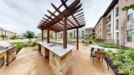 Located at 1400 N Main St in Keller, Texas, this expansive outdoor courtyard is nestled within a multi-story brick residential building complex, offering a well-designed communal outdoor living experience. The centerpiece of the courtyard is a striking pergola structure crafted from rich, dark wood beams, sheltering a built-in outdoor kitchen complete with a granite countertop, stone base, and integrated sink — ideal for outdoor gatherings and entertaining. Surrounding the pergola, multiple seating areas with tables and chairs provide ample space for relaxation and socializing. A generously sized, circular green lawn anchors the heart of the courtyard, bordered by curved walking paths that invite leisurely strolls through the space. Thoughtfully designed landscaping features flowering plants, ornamental grasses, mulched garden beds, and young trees that add color and texture throughout the grounds. The surrounding multi-story brick building presents a classic architectural style with large windows, private balconies, and clean facade detailing. On-site surface parking is conveniently accessible along the building's perimeter. The overall ambiance is welcoming and well-maintained, offering a serene outdoor retreat in the heart of Keller, TX.