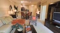 This single-floor residence showcases an open-concept design that connects the living, dining, and kitchen areas into one cohesive, flowing space. The living area is anchored by plush carpeting and features a warm color palette of burnt orange, cream, and neutral tones. A statement zebra-print accent chair with a coral pillow adds a bold, eclectic touch alongside a richly textured rust-colored sofa. A glass-topped coffee table with decorative accessories completes the seating area.

The dining space transitions naturally from the living area and features a high-top table with tall upholstered chairs, ideal for casual dining or entertaining. Just beyond, the kitchen is visible with dark cabinetry, modern appliances including a built-in microwave and refrigerator, and a countertop that doubles as a bar or serving surface.

A built-in shelving and desk area along one wall provides a functional workspace or display nook. Track lighting runs throughout the ceiling, offering ample illumination across all zones. The overall ambiance is warm, inviting, and thoughtfully designed, balancing comfort with a distinct sense of curated style.