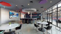 The Hub is a dynamic, single-floor social and amenity space designed with bold contemporary aesthetics and a energetic urban character. At its heart sits a striking circular bar crowned by a large illuminated ring, pendant chandelier, and purple bar stools with gold accents — an eye-catching centerpiece that anchors the entire space. The polished concrete floors reflect the abundant natural light pouring in through floor-to-ceiling glass walls and expansive windows, creating an open, airy atmosphere.

The space is divided into distinct zones that flow seamlessly into one another. A lounge area features plush gray and deep-purple sofas arranged around a modern fireplace set into a bold white-and-black feature wall, with a large mounted TV above. Vibrant, multicolored geometric rugs define the seating zones and inject energy throughout. Decorative laser-cut wall panels and bold graphic wall treatments — including a prominent "Brewerytown" mural — add layers of visual interest.

A casual dining and social zone offers round white tables with contemporary black-and-gold seating, while a game area with a pool table is visible in the background. The entry features striking large-scale abstract wall art on a hexagonal tile floor. The overall ambiance is modern, lively, and social — designed for gathering, relaxing, and entertainment.