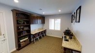 This single-floor study and computer room is thoughtfully designed to accommodate multiple users simultaneously, making it an excellent space for productivity and learning. The room features a long granite-topped built-in desk that runs along one wall, paired with a row of matching dark-stained upper cabinets that provide generous enclosed storage. Several task chairs line the workstation, creating a collaborative workspace suitable for group study or individual tasks. A freestanding dark wood bookcase anchors one side of the room, filled with books, decorative items, and storage baskets, adding both function and warmth to the space. On the opposite side, a second granite-topped workstation accommodates desktop computers and additional equipment, extending the room's functionality as a tech-ready workspace. Recessed lighting throughout the ceiling ensures consistent, well-distributed illumination, while a double-pane window fitted with classic plantation shutters allows natural light to filter in and provides privacy control. The room is finished with neutral blue-gray walls and wall-to-wall carpeting, contributing to a quiet, focused atmosphere. Overall, the space balances practical utility with a refined, classic aesthetic.
