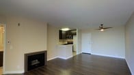 This single-floor space showcases an open-concept layout that seamlessly connects the living area and kitchen. The living room is anchored by a sleek, framed fireplace set against neutral-toned walls, complemented by rich, dark hardwood-style flooring that extends throughout the space, adding warmth and visual continuity.

The kitchen is positioned in the background and features dark espresso cabinetry, stainless steel appliances including a microwave and gas range, and a mosaic tile backsplash that adds a subtle decorative element. A peninsula-style counter defines the kitchen boundary, providing a natural separation from the living area while maintaining the open feel of the layout.

A modern ceiling fan with a sleek, minimalist design is installed overhead, offering both functionality and style. The entry door is positioned adjacent to the kitchen, offering a practical flow upon entry. The smooth, white ceilings and clean, neutral wall tones throughout contribute to a bright, airy atmosphere, making the space feel open and versatile for a variety of furnishing arrangements.