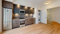 This well-designed one-bedroom, one-bathroom unit in Toronto offers a compact yet functional layout across a single floor. Light hardwood flooring runs throughout, lending a clean and cohesive aesthetic to the entire space.

The kitchen is a standout feature, outfitted with modern two-tone cabinetry — dark upper cabinets paired with warm wood-grain lower cabinets — complemented by recessed lighting and under-cabinet illumination. Stainless steel appliances, including a built-in microwave, electric range, and a wine fridge, add a polished, contemporary touch. A stacked washer and dryer unit is conveniently tucked into an adjacent alcove, providing in-unit laundry.

The living area is bright and open, benefiting from large sliding windows that frame views of the surrounding Toronto streetscape and invite generous natural light. The bedroom is a calm, neutral space with light-toned walls and a large window that continues the theme of natural illumination.

The bathroom is finished with white tile flooring, a full soaker tub with an overhead shower, a wall-mounted vanity with warm wood cabinetry, and recessed ceiling lighting, creating a clean and functional space. A hallway connects all rooms efficiently, rounding out a thoughtfully organized floor plan suited to urban living.