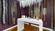This intimate wine room is a thoughtfully designed space that blends bold aesthetics with functional storage. The walls are finished in a rich, deep purple that creates a dramatic backdrop for the extensive floor-to-ceiling wall-mounted wine racks, which line nearly every wall and display a wide variety of bottled wines in an organized, visually striking arrangement. Warm-toned hardwood flooring anchors the space and adds a sense of natural warmth that contrasts beautifully with the vibrant wall color. At the center of the room stands a sleek, high-gloss white serving table, offering a clean and modern surface for wine service and display. A standout design element is the sculptural chandelier overhead, composed of cascading clear loop-shaped forms that catch the light and serve as an artistic focal point. A full-length glass wall and door panel opens the room to an adjacent space, enhancing the sense of openness. Polished 3D lettering spelling "WINE ROOM" is mounted on the glass entry wall, adding a refined and welcoming touch. This space is well-suited for wine storage, tastings, and display, combining functionality with an upscale, curated ambiance throughout.