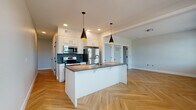 Located at 3600 Johnson Mill Blvd in Johnson, Arkansas, this single-floor unit showcases a cohesive modern design highlighted by striking herringbone-patterned hardwood flooring that flows seamlessly throughout every room. The open-concept kitchen serves as the centerpiece of the space, anchored by a substantial island with a dark countertop, an undermount sink, and a brass-finished faucet. White shaker cabinetry lines the walls, complemented by a teal tile backsplash and a full suite of stainless steel appliances, including a refrigerator, range, and built-in microwave. Sleek black pendant lights with gold interiors hang above the island, adding a contemporary accent. The living area extends naturally from the kitchen, bathed in natural light from a bank of large windows that frame views of surrounding greenery. The bedrooms continue the herringbone flooring theme, each featuring neutral gray walls, natural light from well-placed windows, and ceiling fans for added comfort. One bedroom includes multiple doors with black hardware accents. The bathroom maintains the same design language with a dark countertop vanity, white cabinetry with gold hardware, and herringbone flooring, lending a polished, unified aesthetic throughout the entire unit.