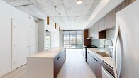 Located in the heart of Salt Lake City, this contemporary 2-bedroom, 1-bathroom residence showcases an industrial-modern aesthetic defined by striking exposed concrete ceilings, light hardwood flooring, and an abundance of natural light. The open-concept kitchen is a standout, featuring rich walnut cabinetry, a soft blue-green subway tile backsplash, stainless steel appliances, and a generously sized waterfall island illuminated by elegant brass pendant lights — ideal for both cooking and entertaining. The living area flows seamlessly toward floor-to-ceiling windows that frame urban views of the surrounding cityscape. The bedrooms are carpeted for comfort and benefit from expansive windows that flood the rooms with natural light and offer additional city views. Each bedroom is complemented by a well-organized closet with built-in shelving and hanging space. The bathroom is thoughtfully designed with light hardwood-style flooring, a warm walnut vanity with a clean white countertop, a large mirror, and a tiled walk-in shower equipped with a fold-down bench and accessibility grab bars. In-unit laundry adds everyday convenience. This residence combines urban sophistication with functional design in a prime downtown Salt Lake City location.