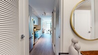 This single-floor residence offers a thoughtfully designed layout that maximizes functionality and style. The entry hallway sets a welcoming tone, featuring warm hardwood-style flooring, a louvered door providing storage access, and a decorative gold-framed mirror that adds a touch of elegance upon arrival.

The galley-style kitchen is efficiently arranged, showcasing light grey flat-panel cabinetry, stone countertops, and a white subway tile backsplash. Stainless steel appliances — including a French door refrigerator, electric range, over-the-range microwave, and dishwasher — provide a cohesive, modern aesthetic. Under-cabinet lighting enhances both functionality and ambiance.

The kitchen flows naturally toward the open living area, which benefits from a large window that draws in generous natural light and offers views of the surrounding streetscape. The living space comfortably accommodates a seating arrangement, making it ideal for relaxing or entertaining.

The bedroom is a calm retreat, featuring textured accent wallpaper behind the bed, a built-in double closet with clean white doors, and soft, neutral tones throughout.

The bathroom is finished with large-format light tile flooring, white subway-tiled tub and shower surround with a decorative niche, and a streamlined vanity with dark countertop detailing.