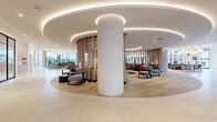 The Ivy presents a meticulously designed lobby and amenity floor that blends modern elegance with organic warmth. The entry is anchored by a dramatic living green wall bearing the Ivy signature, setting a fresh, botanical tone that carries through the space. Large-format light stone tile floors transition seamlessly into wide-plank white oak hardwood as the layout curves gracefully through distinct activity zones.

A defining architectural feature is the sweeping circular lounge at the heart of the space, framed by a cylindrical slatted wood screen and topped with tiered coved ceilings accented by recessed LED lighting. Substantial white columns punctuate the open floor plan, while clusters of contemporary upholstered seating in neutral and warm tones invite gathering. A suspended fireplace adds an intimate focal point to one lounge area.

A row of private workspaces and meeting rooms lines one corridor, enclosed in sleek floor-to-ceiling black-framed glass partitions that maintain visual openness while providing acoustic separation. Natural light pours in through expansive black-framed windows and glass entry doors, offering views of the surrounding exterior. Statement chandeliers with gold hardware provide layered ambient lighting throughout. Curated artwork adorns the walls, and the overall ambiance is polished, refined, and welcoming.