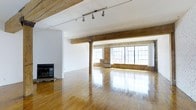 This open-plan loft showcases a striking blend of industrial and rustic character. The expansive main area is defined by its soaring ceiling with exposed wooden beams and a prominent central support column, lending authentic structural charm. Polished hardwood floors run throughout, reflecting abundant natural light that pours in through a row of large, multi-pane industrial windows along the far wall. The windows are framed by exposed brick, adding warmth and texture to the otherwise clean, white-painted walls and ceiling. A fireplace anchors one end of the space, providing a focal point and a sense of warmth. Track lighting mounted along the ceiling beams offers flexible, adjustable illumination to complement the natural light. The layout is wide and open, offering exceptional versatility for a variety of uses, from living and entertaining to creative or professional work. The combination of raw materials — brick, timber, and hardwood — alongside the bright, airy atmosphere creates a dynamic and inviting environment that balances industrial heritage with modern liveability.