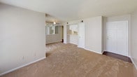 This single-floor, one-bedroom, one-bathroom unit offers a functional and comfortable layout designed to make efficient use of its space. The living area is generously sized, featuring neutral-toned carpet, light beige walls, and a textured ceiling. A bright window allows natural light to fill the room, creating an open and airy feel. A ceiling fan with a light fixture is also present. The living area connects seamlessly to a pass-through kitchen, which is outfitted with white cabinetry, light-colored countertops, a double-basin stainless steel sink, a dishwasher, and a coil-burner electric range — all in white. The kitchen features durable wood-look flooring, providing a practical contrast to the carpeted living spaces. The bedroom is a well-proportioned room with plush carpeting, a window overlooking a green outdoor area, a ceiling fan, and a reach-in closet with sliding doors, offering ample storage. A secondary room or area with mirrored sliding doors provides additional closet or storage space. The bathroom includes a shower-tub combination, a toilet, overhead cabinetry for storage, and a vanity. The overall ambiance of the unit is clean, neutral, and move-in ready.