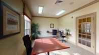 This single-floor business center offers a thoughtfully designed workspace that balances functionality with a warm, professional aesthetic. The room is finished with soft cream-toned walls accented by rich wood crown molding, and neutral-toned carpeting throughout that contributes to a calm, focused atmosphere.

The space is organized to support multiple users simultaneously. Along the walls, built-in rich mahogany-finished desks provide ample workstation space, each equipped with desktop computers, ergonomic mesh-back chairs, and a shared printer, making the area well-suited for everyday computing tasks. A standalone central table with a polished wood surface offers additional meeting or workspace flexibility.

Natural light flows generously through large multi-pane windows dressed with patterned valances in warm earth tones, offering views of the surrounding outdoor landscape with seasonal foliage visible. Overhead, recessed fluorescent lighting ensures consistent illumination throughout the room.

Decorative touches include framed artwork on the walls and lush potted plants positioned near the windows, adding a touch of greenery and life to the environment. Access to the space is provided through a classic French door with glass panels, lending an open and inviting feel. The overall design reflects a practical yet comfortable environment conducive to work and productivity.