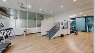 Located at 50 Park Row West in Providence, Rhode Island, this single-floor fitness and aquatic facility offers a generous, well-appointed space designed to support a comprehensive wellness experience. The exercise room spans a substantial area laid with light hardwood-style flooring and features a drop ceiling with recessed lighting and ceiling fans that maintain a bright, comfortable atmosphere throughout. The fitness area is divided into distinct zones, including a cardio section equipped with treadmills, stationary bikes, and rowing machines, as well as a strength training area outfitted with cable machines, free weights, a weight bench, and functional training equipment such as a RAX multi-purpose rig. Teal accent walls, wall-mounted monitors, and full-length mirrors enhance the modern aesthetic while keeping the space practical and motivating. A staircase with metal railings connects sections of the facility, and frosted glass partition walls add a refined architectural detail near the entry area. The indoor pool is a standout feature, boasting calm, clear water and a surrounding tile deck. Large floor-to-ceiling windows and glass doors flood the pool area with natural light and offer views of the outdoors. An accessibility lift is available at poolside, ensuring inclusive use. Seating areas adjacent to the pool provide a place to relax, completing this well-rounded facility.