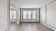 This 2-bedroom, 1-bathroom unit is laid out across a single floor, offering a well-organized and efficient use of space. The living areas are immediately distinguished by elegant crown molding, decorative wall trim panels, and wood-style plank flooring in a warm neutral tone that runs throughout the entire unit. Large windows fitted with horizontal blinds flood the living rooms with natural light, creating a bright and airy atmosphere. The open archway connecting the two living areas adds architectural character and a sense of flow.

The kitchen is outfitted with white shaker-style cabinetry, granite countertops featuring a classic white and grey pattern, a stainless steel sink with a modern faucet, and stainless steel appliances including a dishwasher. Open upper shelving provides additional storage and display space.

The primary bedroom is a comfortable size, featuring crown molding, wood-style flooring, and a window that brings in natural light. The second bedroom offers a cozier footprint with its own window and a distinctive painted white brick accent wall that adds texture and character.

The bathroom includes a soaking tub, a white vanity with under-cabinet storage, a wall-mounted mirror, and a window with frosted glass for privacy. Wood-style flooring ties this room to the rest of the unit cohesively.