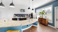Located in Hickory, NC, this single-floor community amenity space offers a thoughtfully designed layout that blends social, recreational, and functional areas seamlessly. The open-concept kitchen and living area serves as the heart of the space, featuring a striking curved granite-topped island with a bold teal base, dark cabinetry with granite countertops, stainless steel appliances, and stylish pendant lighting. Large windows flood the kitchen with natural light and offer views of the surrounding greenery. The living areas are finished with crown molding, recessed lighting, and a sophisticated color palette of teal accent walls, crisp white trim, and warm wood-look flooring. A generously sized lounge area features a tufted leather Chesterfield sofa, patterned carpet, and a large wall-mounted television, making it ideal for relaxing or gathering. The game room section includes a full-size pool table and a dartboard, providing ample entertainment options. A dedicated package room houses smart parcel lockers for convenient, secure deliveries. Throughout the space, abundant windows invite natural light and outdoor views, contributing to a bright, welcoming ambiance that encourages community interaction and comfort.