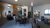 This well-appointed single-floor space offers an inviting open-concept layout that seamlessly blends a dining area and lounge into a cohesive, functional environment. The dining area is anchored by a round dark-wood table surrounded by elegant dark-framed chairs with black upholstered seating, set atop a patterned area rug that adds texture and visual interest underfoot. Overhead, modern pendant lights provide warm, focused illumination that enhances the dining experience. The adjacent lounge area features a comfortable sofa and is highlighted by a fireplace framed in warm wood tones and a rich burnt-orange accent wall, creating a welcoming focal point. Decorative wall art, including a striking multi-panel vertical piece and an eclectic metallic wall sculpture, add personality and artistic flair throughout the space. A large decorative tree and wall-mounted sconce lighting contribute to the space's layered ambiance. Neutral gray walls and carpeted floors run throughout, lending a calm, unified aesthetic. Architectural archways define transitions between zones while maintaining an open, airy feel. The overall design strikes a balance between modern sophistication and comfortable functionality, making it well-suited for gathering and socializing.