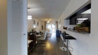 This single-floor residence is designed with an open-concept layout that seamlessly connects the kitchen, dining, and living areas into one cohesive living space. Rich, dark hardwood flooring runs throughout, lending warmth and a sense of continuity to the interior.

The kitchen is positioned along one side of the main living area and features dark cabinetry, stone countertops, and a breakfast bar with sleek modern barstools, making it ideal for casual dining or entertaining. Pendant lighting above the bar adds a stylish focal point.

Adjacent to the kitchen, the dining area accommodates a round table with contemporary white chairs, illuminated by an overhead pendant light that enhances the intimate atmosphere of the space.

The living room anchors the far end of the open floor plan, centered around a ceiling fan and furnished with comfortable seating. A large sliding glass door floods the space with natural light and opens onto a balcony, offering views of lush greenery beyond.

Throughout the space, neutral wall tones complement the dark flooring and cabinetry, creating a balanced and modern aesthetic. The textured ceiling and thoughtful lighting choices add dimension and character, making this a practical yet stylish single-floor home.