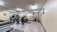 This exercise room at 17830 Sherman Way in the Reseda neighborhood of Los Angeles, CA offers a dedicated fitness space efficiently arranged on a single floor. The room features light wood-look flooring and warm yellow-toned walls, creating a bright and welcoming atmosphere. Overhead fluorescent lighting ensures the space is well-illuminated throughout, complemented by a wall-mounted air conditioning unit that maintains a comfortable workout environment. A large wall mirror runs along one side of the room, visually expanding the space and allowing users to monitor their form during exercise. The fitness area is equipped with multiple Star Trac treadmills, a stationary upright bike, a recumbent bike, an elliptical trainer, and a multi-function cable strength machine, providing a well-rounded selection of cardio and resistance training options. A textured ceiling adds a classic architectural touch, while dark baseboards provide a clean contrast to the light flooring. The room is accessed through a solid entry door with an automatic door closer, ensuring privacy and security. Overall, this exercise room delivers a practical and functional fitness experience, making it a convenient amenity for residents or guests at this Los Angeles location.
