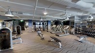 This spacious fitness center is designed to accommodate a full range of workout routines within a single open floor layout. The facility features an industrial-inspired ceiling with exposed white-painted pipes and ductwork, complemented by pendant-style industrial lighting that provides even, functional illumination throughout the space. The flooring is covered in a patterned carpet with subtle wave detailing, adding texture and comfort underfoot.

The cardio section is prominently featured, housing a row of modern treadmills equipped with individual digital display screens, offering users an interactive workout experience. Alongside the treadmills, a rowing machine adds variety to the cardio options available. The strength training area is well-appointed with multiple resistance machines and free weight benches arranged to allow ample space between equipment.

Mirrored walls line the perimeter, visually expanding the space and allowing users to monitor their form during exercise. Structural columns wrapped in a sleek blue-toned finish add a polished, contemporary aesthetic to the room. The overall ambiance is clean, organized, and professional, creating an environment well-suited for focused, efficient training sessions.