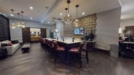This elegantly designed single-floor communal space blends modern sophistication with warm, inviting details across several distinct zones. At its heart is a generously sized kitchen and bar area anchored by a large island topped with light granite countertops and lined with dark wood bar stools upholstered in rich burgundy leather. A dramatic textured accent wall in woven wood tones serves as a striking backdrop, complemented by a wall-mounted flat-screen TV and a suite of stainless steel appliances beneath a sleek industrial range hood. Overhead, sculptural gold pendant lights add a refined focal point.

Adjacent to the kitchen, a comfortable lounge area features a linear gas fireplace set into a clean, horizontal-shelved wall, accompanied by plush gray seating, a chaise, and a statement woven pendant light fixture. Clerestory windows allow natural light to filter in, enhancing the warm ambiance.

A separate seating nook showcases a bold grid-pane glass wall that frames views into an adjacent space, paired with a classic dark wood sliding barn door. Rich hardwood flooring flows throughout, tying each area together seamlessly. A billiards area with deep red wingback chairs and framed wall art rounds out the space, offering a relaxed retreat ideal for socializing and recreation.