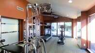 This fitness center is designed to support a comprehensive workout experience within a compact, efficiently organized space. The room features warm terracotta-toned accent walls paired with light wood-style flooring, creating an energizing yet welcoming atmosphere. A prominent multi-station cable machine with chrome pulleys and bars anchors the space, offering a wide range of strength training options. Cardio equipment, including a treadmill and an elliptical trainer, is positioned along the perimeter and oriented toward large floor-to-ceiling windows. These expansive windows flood the room with natural light and provide a pleasant view of mature trees and outdoor greenery, making workouts feel connected to the outdoors. Recessed ceiling lighting supplements the natural light, ensuring the space is well-illuminated at all times. Wall-mounted mirrors enhance the sense of depth and allow for proper form checks during exercise. A mounted display screen is also present, offering entertainment or instructional content during workouts. The single-floor layout keeps everything accessible and within reach, making this fitness center a practical and inviting facility for daily use.