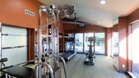 This fitness center is designed to support a comprehensive workout experience within a compact, efficiently organized space. The room features warm terracotta-toned accent walls paired with light wood-style flooring, creating an energizing yet welcoming atmosphere. A prominent multi-station cable machine with chrome pulleys and bars anchors the space, offering a wide range of strength training options. Cardio equipment, including a treadmill and an elliptical trainer, is positioned along the perimeter and oriented toward large floor-to-ceiling windows. These expansive windows flood the room with natural light and provide a pleasant view of mature trees and outdoor greenery, making workouts feel connected to the outdoors. Recessed ceiling lighting supplements the natural light, ensuring the space is well-illuminated at all times. Wall-mounted mirrors enhance the sense of depth and allow for proper form checks during exercise. A mounted display screen is also present, offering entertainment or instructional content during workouts. The single-floor layout keeps everything accessible and within reach, making this fitness center a practical and inviting facility for daily use.
