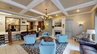 This spacious single-floor facility presents a thoughtfully designed common area that combines elegance with comfort. The centerpiece of the space is a grand lounge featuring a striking coffered ceiling adorned with crisp white molding, from which a classic wrought-iron chandelier with warm-toned shades hangs as a focal point. A decorative fireplace flanked by powder-blue cabinetry with cross-panel detailing anchors one wall, topped by a large landscape painting that adds an artistic touch to the inviting atmosphere.

Seating throughout the lounge is carefully arranged to encourage conversation and relaxation, with a rich brown leather sofa and coordinating powder-blue upholstered armchairs gathered around a round wooden coffee table atop a bold patterned area rug. Light tile flooring runs throughout, contributing to the bright, airy feel of the space.

Large windows draped in elegant gray curtains allow natural light to pour in, offering outdoor views that further enhance the welcoming ambiance. A carpeted staircase with decorative patterned runner and dark wood banisters connects to additional levels, alongside elevator access. Wide, carpeted corridors lined with warm wood-toned doors extend from the main lounge, providing a well-organized and accessible flow throughout the entire facility.