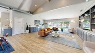 Located at 6312 Birdcage St in Citrus Heights, CA, this thoughtfully designed single-floor space offers a generous, open-concept layout that seamlessly integrates a living room and dining area. The interior is anchored by striking vaulted ceilings with exposed ductwork that lends an airy, loft-inspired character throughout. Light wood-look flooring flows consistently across the entire space, complementing the neutral, contemporary color palette.

The living area is a standout, featuring a custom built-in media wall with gray shaker-style cabinetry, open display shelving, and ample storage. Large sliding glass doors span the exterior wall, flooding the room with natural light and offering direct views of a lush green outdoor area. A dedicated coffee bar station with additional cabinetry adds a practical and inviting touch.

The dining area is framed by a full glass partition wall with sliding doors, creating a semi-private yet visually connected space. A modern pendant light fixture anchors the dining table, while French doors provide a second point of access to the outdoors. Additional seating nooks with built-in counters offer versatile, informal gathering spots. Together, these elements create a bright, welcoming, and highly functional communal environment.