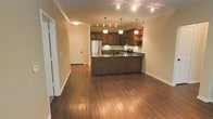 This single-floor space showcases an open-concept layout that seamlessly connects the kitchen and living areas, creating a spacious and functional environment. Rich hardwood flooring flows throughout, adding warmth and visual continuity to the space.

The kitchen is a standout feature, anchored by a large peninsula with a dark stone countertop that provides ample workspace and doubles as a casual dining or entertaining area. Warm-toned wooden cabinetry lines the walls, offering generous storage, while stainless steel appliances — including a refrigerator and microwave — add a modern touch. Pendant lighting hangs above the peninsula, complementing the track lighting overhead to create a well-lit and inviting atmosphere.

Crown molding runs along the ceiling, contributing a refined architectural detail that elevates the overall aesthetic. The open sightlines between the kitchen and adjacent living area make the space feel expansive and airy. Neutral wall tones serve as a versatile backdrop, allowing the natural warmth of the wood elements to take center stage. The overall ambiance is welcoming, combining classic design details with contemporary functionality.