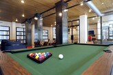 This single-floor community amenity space masterfully blends industrial and modern design elements into a cohesive, inviting environment. Exposed red brick walls, warm wood plank ceilings, and polished concrete floors establish a raw yet refined aesthetic throughout. Bold steel structural columns and visible ductwork add to the industrial character, while large steel-framed windows flood the space with abundant natural light and offer views of the surrounding greenery.

The open layout is divided into distinct functional zones that flow seamlessly into one another. A spacious lounge area features comfortable striped armchairs, leather seating, and a wall-mounted television, creating an ideal spot for relaxation or casual conversation. Adjacent to it, a billiards table anchors a game area illuminated by an Edison-bulb chandelier, completing the lively atmosphere.

A standout coffee and refreshment bar showcases a curved dark-topped island with industrial-style wood barstools, set against a shimmering metallic tile backsplash. Open wood shelving styled with plants and artwork adds warmth to the bar area, which is equipped with a professional coffee machine and sink cabinetry. Steel-framed glass partition walls elegantly define spaces without sacrificing openness. A staircase with a sleek metal cable railing provides vertical access, completing this thoughtfully designed communal retreat.