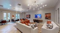 This single-floor communal space is thoughtfully designed to offer both comfort and sophistication. The main lounge area is anchored by a large wall-mounted flat-screen television and a striking live-edge wood console beneath it, paired with plush white sofas and vibrant burnt-orange accent chairs arranged atop a layered area rug over light tile flooring. A sculptural multi-arm chandelier and recessed lighting illuminate the room, which is finished with crisp crown molding throughout.

Curated contemporary artwork adorns the walls, adding warmth and visual interest to the neutral gray-and-white color palette. A glass-top coffee table with geometric legs serves as a central focal point in the lounge seating area.

Adjacent to the main lounge, a dining nook features a round table with leather chairs and benefits from natural light streaming through French doors that overlook an outdoor pool area, offering a bright and inviting setting.

The kitchenette is well-equipped for entertaining, featuring granite countertops, white cabinetry, a stainless steel sink, a beverage refrigerator, and a textured stacked-stone backsplash that adds an upscale architectural detail. Stone tile flooring in this area complements the overall refined aesthetic of the space, making it both functional and visually cohesive.