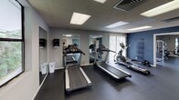 Located at 2601 N John B Dennis Hwy in Kingsport, Tennessee, this fitness center offers a comprehensive workout facility across a single floor. The space is designed to accommodate a wide range of exercise needs, with distinct zones dedicated to cardiovascular training and strength work.

The cardio area is well-equipped with multiple treadmills, elliptical machines, and stationary bikes arranged in an open layout. Large windows throughout the facility invite abundant natural light and provide views of the surrounding greenery, creating an energizing atmosphere. Full-length wall mirrors enhance the sense of space and allow users to monitor their form during workouts.

The strength training zone features a multi-station cable machine, free weights, dumbbells, weight benches, a stability ball, and additional resistance equipment. Wall-mounted cable attachments provide versatility for a variety of exercises. A ceiling fan aids with airflow in this area, and a sliding glass door offers access to an adjacent outdoor space.

Underfoot, durable dark rubber flooring covers the entire facility, providing both safety and comfort. The drop ceiling features recessed panel lighting that ensures the space is brightly and evenly lit throughout. Sanitation stations are conveniently placed within the facility. The entrance area is clean and open, with posted fitness center rules and direct access to the main workout floor, making navigation intuitive for all users.