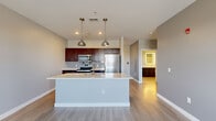 Located at 20 Ellen Polimeni Blvd in Canandaigua, NY, this well-appointed one-bedroom, one-bathroom residence offers a thoughtfully designed layout across a single, generously sized floor. The open-concept kitchen and living area serves as the heart of the home, where rich dark-cabinetry kitchen cabinets provide striking contrast against crisp white quartz countertops. A large central island with an integrated sink and pendant lighting above creates a natural gathering point and seamlessly connects the kitchen to the living space. Stainless steel appliances, including a refrigerator, range, and built-in microwave, complete the kitchen's modern aesthetic. Light wood-tone flooring flows throughout the main living areas, complementing the soft gray walls and white trim. The spacious living room benefits from multiple windows that draw in abundant natural light and provide outdoor views, while a glass-paneled door leads to an exterior access point. The well-proportioned primary bedroom features plush carpeting, a window with natural light, and direct access to the main hallway. The bathroom is finished with a dark vanity, white quartz countertop, an illuminated mirror, and a full tub-shower combination with brushed nickel fixtures and tile flooring, completing this clean and contemporary residence.