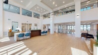 Located at 2900 W Hamlin Rd in Rochester Hills, Michigan, this sophisticated two-story commercial space offers a striking blend of modern design and functional versatility. The ground floor makes an immediate impression with its soaring double-height atrium, anchored by light-toned hardwood flooring that flows seamlessly throughout. Oversized floor-to-ceiling windows and glass-paneled entry doors flood the space with abundant natural light, while dramatic circular pendant light fixtures add a refined architectural focal point overhead. A built-in reception desk with seating creates a polished welcome area, complemented by an open conference table ideal for collaborative gatherings. A vibrant living moss wall installation adds a bold organic element, enhancing the clean white palette of the interior. Glass-enclosed private offices provide visual connectivity while maintaining acoustic separation. The upper floor, accessible via a contemporary staircase with cable railings, features an open mezzanine overlooking the atrium below, along with additional private offices and meeting areas. Intimate nook seating areas with curated artwork offer quiet retreat spaces. A well-appointed utility room with textured wall paneling, modern cabinetry, and exterior access rounds out the amenities. Throughout, the space balances openness and privacy with a refined, professional aesthetic.
