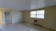 This single-floor space presents a clean, utilitarian layout with a no-frills design suited for practical use. The main room is defined by its distinctive cream-colored square tile wall paneling, which lines the walls from floor to ceiling and gives the space a retro, institutional character. The flooring is composed of vinyl tile in a light neutral tone, contributing to the room's understated, easy-to-maintain aesthetic. A textured ceiling adds a subtle visual detail overhead. Natural light enters generously through a large double-pane window fitted with horizontal blinds, offering views of surrounding greenery and creating a bright, airy atmosphere. Climate control is handled by a wall-mounted window air conditioning unit paired with an under-window baseboard heating system, ensuring year-round comfort. The room is currently open and unfurnished, making its full spatial potential clearly visible. The overall ambiance is functional and bright, offering a blank canvas that could serve a variety of purposes. The straightforward design and durable surface materials suggest a space built with longevity and ease of maintenance in mind.