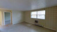 This single-floor space presents a clean, utilitarian layout with a no-frills design suited for practical use. The main room is defined by its distinctive cream-colored square tile wall paneling, which lines the walls from floor to ceiling and gives the space a retro, institutional character. The flooring is composed of vinyl tile in a light neutral tone, contributing to the room's understated, easy-to-maintain aesthetic. A textured ceiling adds a subtle visual detail overhead. Natural light enters generously through a large double-pane window fitted with horizontal blinds, offering views of surrounding greenery and creating a bright, airy atmosphere. Climate control is handled by a wall-mounted window air conditioning unit paired with an under-window baseboard heating system, ensuring year-round comfort. The room is currently open and unfurnished, making its full spatial potential clearly visible. The overall ambiance is functional and bright, offering a blank canvas that could serve a variety of purposes. The straightforward design and durable surface materials suggest a space built with longevity and ease of maintenance in mind.