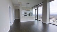 This contemporary high-rise unit is designed with an open-concept layout that seamlessly connects the living, dining, and kitchen areas into one cohesive and functional space. Dark hardwood floors run throughout, providing a rich contrast against the light grey walls and crisp white finishes. The modern kitchen features white cabinetry, a peninsula with a built-in sink, stainless steel appliances including a refrigerator with ice dispenser, microwave, and range, all topped with light countertops. A stylish multi-bulb pendant light fixture hangs above the peninsula, adding a contemporary focal point. The living area is bathed in natural light, courtesy of expansive floor-to-ceiling windows that frame breathtaking panoramic views of the surrounding landscape and skyline. A sliding glass door provides direct access to a private balcony, extending the living space outdoors. A hallway leads to additional rooms, maintaining a clean and organized flow throughout the unit. The overall ambiance is modern, airy, and elevated, with high-quality finishes and an abundance of natural light creating a refined and comfortable living environment well-suited for those who appreciate contemporary urban design.