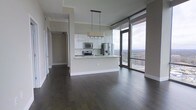 This contemporary high-rise unit is designed with an open-concept layout that seamlessly connects the living, dining, and kitchen areas into one cohesive and functional space. Dark hardwood floors run throughout, providing a rich contrast against the light grey walls and crisp white finishes. The modern kitchen features white cabinetry, a peninsula with a built-in sink, stainless steel appliances including a refrigerator with ice dispenser, microwave, and range, all topped with light countertops. A stylish multi-bulb pendant light fixture hangs above the peninsula, adding a contemporary focal point. The living area is bathed in natural light, courtesy of expansive floor-to-ceiling windows that frame breathtaking panoramic views of the surrounding landscape and skyline. A sliding glass door provides direct access to a private balcony, extending the living space outdoors. A hallway leads to additional rooms, maintaining a clean and organized flow throughout the unit. The overall ambiance is modern, airy, and elevated, with high-quality finishes and an abundance of natural light creating a refined and comfortable living environment well-suited for those who appreciate contemporary urban design.