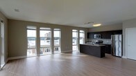 This single-floor space showcases a thoughtfully designed open-concept layout that seamlessly integrates the living area and kitchen. Large floor-to-ceiling windows line two walls, flooding the interior with natural light and providing expansive views of the surrounding urban skyline. The light hardwood flooring extends throughout the space, contributing to a clean, modern aesthetic.

The kitchen is a standout feature, equipped with dark espresso cabinetry that contrasts elegantly with the light tones of the surrounding space. A central island with a granite countertop serves as both a functional workspace and a natural dividing element between the kitchen and living area. Stainless steel appliances, including a microwave and range, are seamlessly integrated into the cabinetry. A track lighting fixture above the kitchen provides focused illumination, complementing the abundant natural light throughout.

The open living area is generous in scale, offering flexibility for various furniture arrangements. Neutral-toned walls and white ceilings enhance the brightness and sense of openness, while the overall design maintains a contemporary, minimalist character throughout.