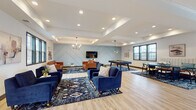 Located at 3552–3622 NE Akin Dr in Lee's Summit, Missouri, this single-floor community space is designed to foster social interaction and relaxation through a thoughtfully curated, modern interior. The open layout is anchored by light hardwood-style flooring that runs throughout, creating a cohesive and airy feel across the entire space.

The main lounge area is generously sized and defined by tray ceilings with recessed lighting and contemporary pendant chandeliers that add warmth and visual interest. A signature accent wall, painted in a soft slate blue and adorned with a geometric chevron-pattern millwork design, serves as a striking focal point. Mounted below it, a flat-screen television is flanked by two tan leather sofas arranged over a bold patterned area rug, creating an inviting media lounge. Navy blue velvet armchairs and sofas populate additional seating zones, offering flexible arrangements for gatherings of various sizes.

A game area with a billiards table is positioned to one side, while a dedicated kitchenette provides practical amenities including dark Shaker-style cabinetry, white hexagonal tile backsplash, stainless steel appliances, and light countertops. A glass-paneled door in the kitchenette opens to an outdoor area, allowing natural light to fill the space. Large windows throughout further enhance the bright, welcoming atmosphere.