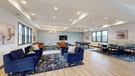 Located at 3552–3622 NE Akin Dr in Lee's Summit, Missouri, this single-floor community space is designed to foster social interaction and relaxation through a thoughtfully curated, modern interior. The open layout is anchored by light hardwood-style flooring that runs throughout, creating a cohesive and airy feel across the entire space.

The main lounge area is generously sized and defined by tray ceilings with recessed lighting and contemporary pendant chandeliers that add warmth and visual interest. A signature accent wall, painted in a soft slate blue and adorned with a geometric chevron-pattern millwork design, serves as a striking focal point. Mounted below it, a flat-screen television is flanked by two tan leather sofas arranged over a bold patterned area rug, creating an inviting media lounge. Navy blue velvet armchairs and sofas populate additional seating zones, offering flexible arrangements for gatherings of various sizes.

A game area with a billiards table is positioned to one side, while a dedicated kitchenette provides practical amenities including dark Shaker-style cabinetry, white hexagonal tile backsplash, stainless steel appliances, and light countertops. A glass-paneled door in the kitchenette opens to an outdoor area, allowing natural light to fill the space. Large windows throughout further enhance the bright, welcoming atmosphere.