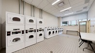Located at 695 Atlantic St in Stamford, Connecticut, this shared laundry facility offers a well-organized and efficiently designed space across a single floor. The room is outfitted with a generous bank of front-loading washing machines aligned along one wall, complemented by stacked dryer units positioned nearby — providing substantial combined laundry capacity for building residents. The space features clean white square floor tiles throughout, contributing to a hygienic and bright atmosphere. Overhead, a dropped ceiling with flush-mounted fluorescent lighting ensures consistent, even illumination throughout the room. Natural light filters in through windows fitted with roller shades, offering views of surrounding greenery. A folding table and seating are conveniently provided, giving users a dedicated surface for sorting and folding laundry. The walls are finished in neutral tones, maintaining a clean and functional aesthetic. A utility station and payment kiosk are also present, streamlining the laundry experience. Access is provided through a glass-paneled door, and the layout includes an entryway corridor with white tiled walls that leads into the main laundry area. Overall, the facility is thoughtfully arranged to maximize usability while maintaining a tidy, utilitarian environment.