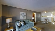 This single-floor space features a welcoming living area finished in cool grey tones, creating a calm and cohesive aesthetic. The living room is centered around a teal tufted loveseat accented with floral throw pillows, complemented by a bold yellow coffee table that adds a vibrant contrast to the neutral palette. A framed floral canvas artwork adorns the grey accent wall, enhancing the room's decorative character. The textured ceiling adds architectural interest overhead, while carpeted flooring provides warmth underfoot. A doorway visible from the living area leads to an adjoining room, suggesting a functional layout with well-connected spaces. To one side, a glimpse of a dining area is visible, featuring white chairs, indicating a dedicated space for meals. A table lamp provides warm ambient lighting, contributing to the overall inviting atmosphere. The space is served by ceiling-mounted HVAC vents, ensuring year-round comfort. Overall, the unit presents a thoughtfully arranged interior with a harmonious blend of cool and warm tones, practical layout, and comfortable furnishings suited for everyday living.