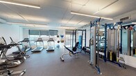 This fitness center offers a comprehensive workout environment designed to support a wide range of exercise routines. The space is organized across a single floor and is divided into distinct functional zones, each dedicated to a specific type of training.

The cardio area is positioned along a windowed wall fitted with roller shades, allowing ample natural light to filter in while maintaining privacy. Multiple treadmills face the windows, providing an open and airy feel during use. A collection of stability and medicine balls on a standing rack adds versatility to this zone.

Adjacent to the cardio area, a dedicated strength and resistance training section features a multi-functional cable and weight machine, a flat utility bench, and a full rack of dumbbells arranged along a mirrored wall. The mirrors enhance the sense of space and allow users to monitor their form during exercise.

A separate section of the fitness center houses additional cardio and low-impact equipment, including elliptical trainers and a stationary cycle. This area features a wall-mounted flat-screen television and is accented by a dark, textured feature wall, giving it a distinct and modern aesthetic.

Throughout, rubber flooring provides durability and safety, while suspended linear LED lighting ensures consistent, bright illumination. The clean white walls and modern finishes contribute to a professional and motivating atmosphere.