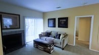 This single-floor residence offers a warm and functional living environment designed for everyday comfort. The living room serves as the central gathering space, featuring soft wall-to-wall carpeting underfoot and walls painted in a soft, warm yellow tone that lends a welcoming ambiance to the room. A large sliding glass door fitted with vertical blinds allows an abundance of natural light to fill the space while providing access to the outdoors. The living area is thoughtfully arranged with a plush sofa accented by decorative throw pillows and anchored by a distinctive wooden trunk-style coffee table, adding a touch of rustic charm. Two framed wall art pieces add a finishing touch to the aesthetic. A ceiling vent indicates a central air system for year-round climate control. The open doorway visible from the living room suggests a seamlessly connected floor plan, allowing easy flow between the living spaces. Overall, the residence presents a cozy, practical, and well-lit interior well-suited for comfortable everyday living.