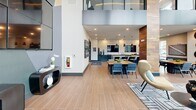 Located at 5580 Farrington Rd in Durham, NC, this contemporary amenity facility offers a sophisticated blend of social and fitness spaces spread across a single expansive floor. The architectural design is immediately striking, with soaring double-height ceilings, floor-to-ceiling windows, and reclaimed wood accent columns that infuse warmth into the modern aesthetic.

The welcoming entrance transitions into an open lounge area featuring stylish furnishings, including a plush purple sectional, sculptural accent chairs, and geometric area rugs laid over wood-look tile flooring. A communal dining and kitchen area anchors the social zone, complete with a large island, pendant lighting, terrazzo-inspired backsplash, dark cabinetry, and mounted flat-screen TVs — ideal for gatherings and events.

The fitness center is a standout feature, generously sized and divided into distinct zones. Cardio equipment including treadmills, ellipticals, and stair climbers line the perimeter beneath expansive windows offering views of the surrounding natural landscape. Strength training areas feature free weights, cable machines, resistance equipment, and functional training rigs. A dramatic geometric mosaic accent wall adds visual interest to the workout space.

An upper mezzanine level, visible through glass railings, adds architectural depth. Throughout the facility, recessed lighting, rich wood paneling, and curated artwork contribute to a polished, upscale ambiance.